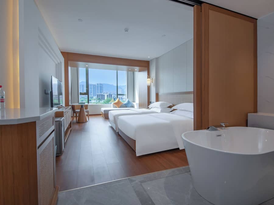 A modern hotel room with a large bed, a separate bathroom with a freestanding bathtub, a desk with a chair, and a large window offering a city view.