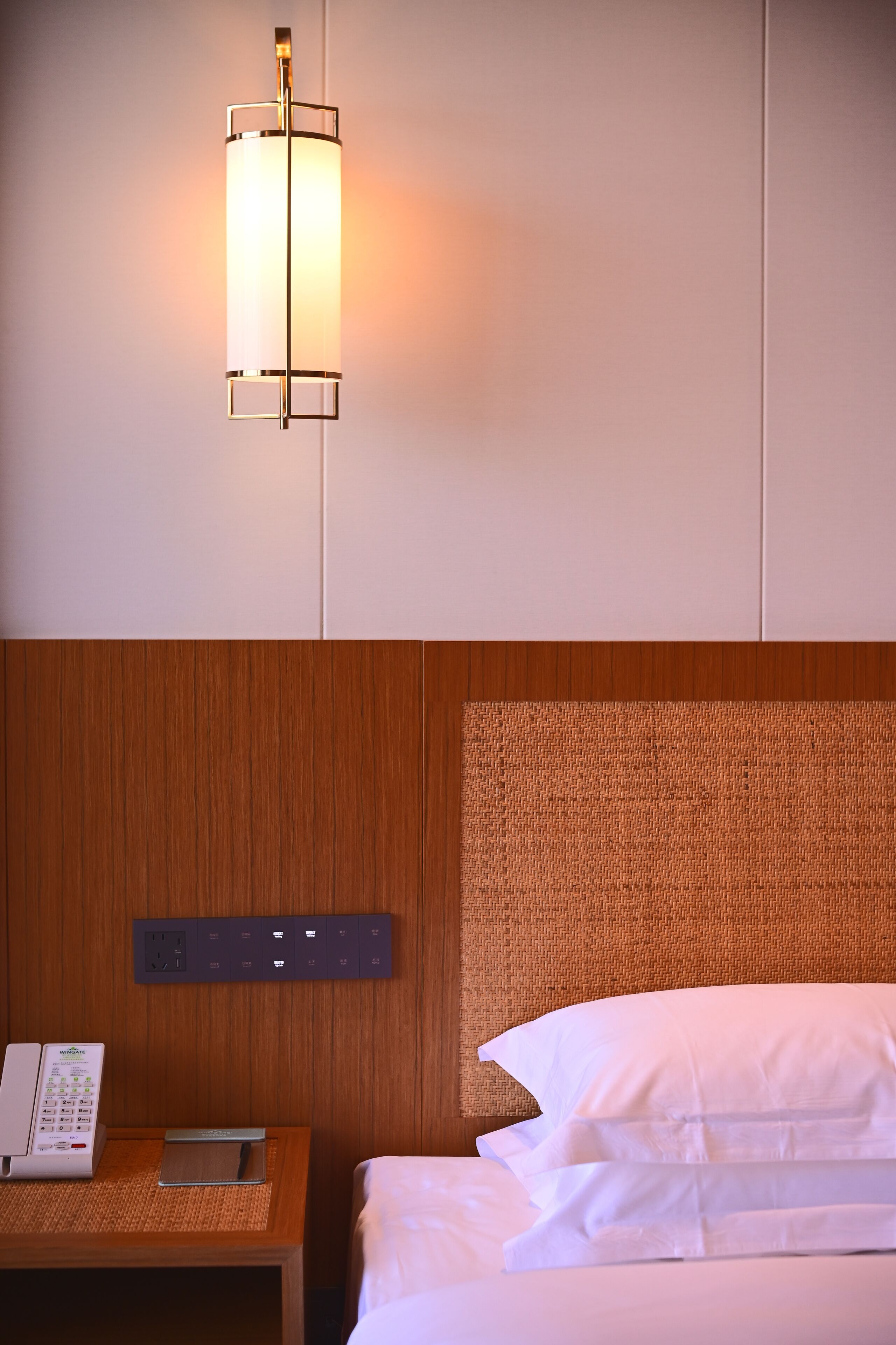 A hotel room with a wooden headboard, a bedside table with a phone and a small electronic device, and a wall-mounted light fixture.