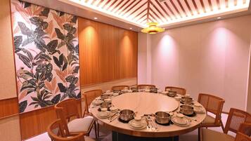 Un salón de conferencias con una mesa redonda, sillas de madera y una pared decorativa con hojas tropicales.