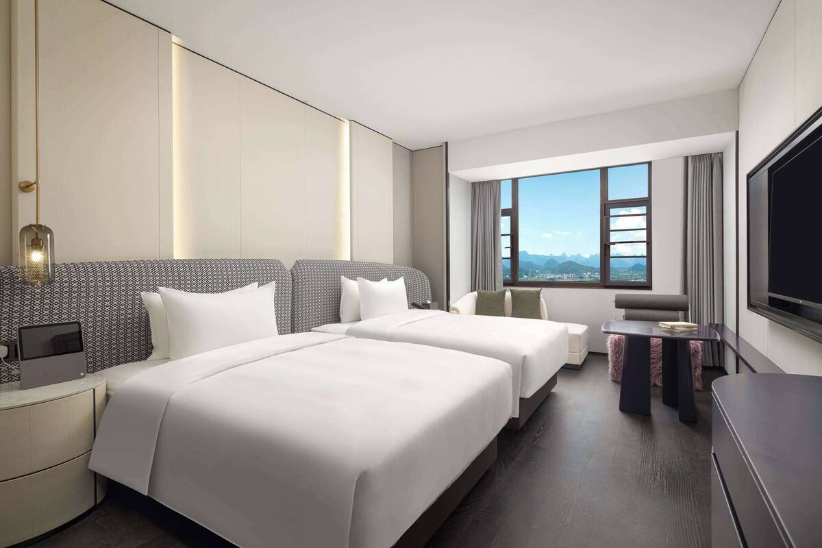 Ein Hotelzimmer mit zwei Betten, einem Schreibtisch, einem Stuhl, einem Fernseher und einem großen Fenster mit Blick auf die Berge.
