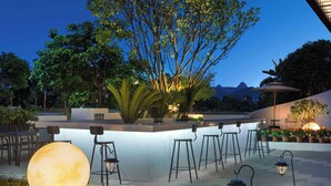 Uma área de bar ao ar livre com disposição de assentos, uma grande estrutura iluminada semelhante à lua, e um cenário de árvores e montanhas.