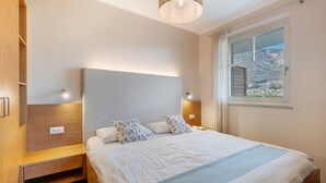 Un dormitorio con cama, lámparas de noche, mesita de noche, armario de madera y ventana con vistas a la montaña.