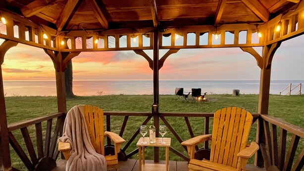 A covered outdoor seating area with two wooden Adirondack chairs, a table with a glass of wine, and a view of the ocean at sunset. - Relax on the Lake - 180 degree Views (North East)