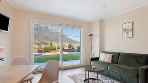 A living room with a green sofa, a small round table, a TV mounted on the wall, and a large sliding glass door leading to a garden.