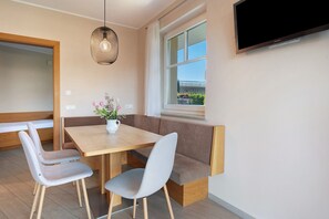 A modern dining area with a wooden table, two grey chairs, a bench, a hanging light fixture, and a TV mounted on the wall.