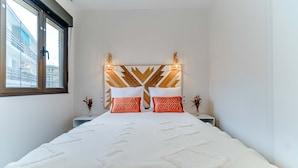 A bedroom with a large bed, a geometric headboard, two bedside tables, and a window with a view of buildings.