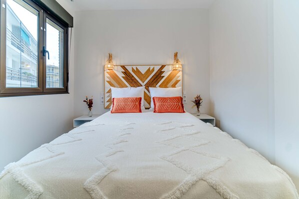 A bedroom with a large bed, a geometric headboard, two bedside tables, and a window with a view of buildings.