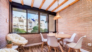 A modern dining area with a wooden table and white chairs, a cushioned chair, and a large window offering a view of buildings and trees.