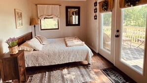 A bedroom with a bed, a bedside table, a lamp, a window with curtains, a mirror, and a view of the outside deck.