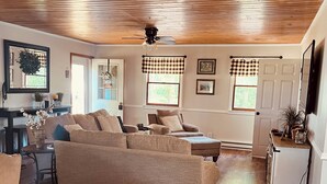 A spacious living room with a large beige sectional sofa, wooden flooring, a ceiling fan, and a kitchen area with a refrigerator.