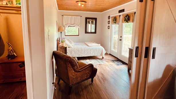 A room with a wooden floor, a bed with a white bedspread, a brown armchair, and a sliding door leading to another room.