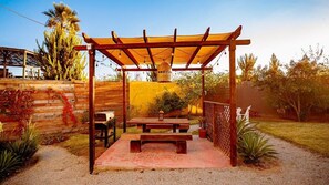 Eine Holz-Pergola mit Tisch und Stühlen darunter, umgeben von einem Garten mit verschiedenen Pflanzen und einem Zaun.