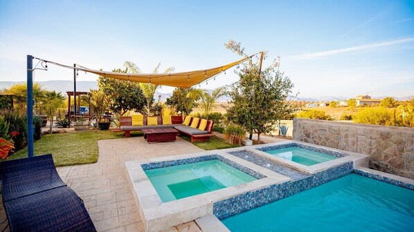 Un patio trasero con piscina, una zona de estar con cojines y una pérgola.