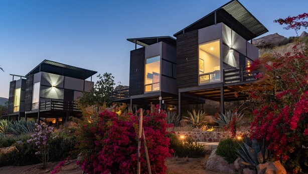 A modern two-story house with large windows and a balcony, surrounded by desert vegetation.