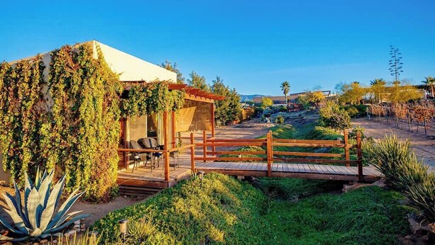 Una cabaña rústica con terraza de madera y zona de estar cubierta, rodeada de vegetación exuberante y un cielo azul despejado.