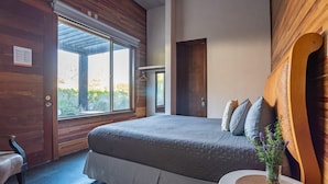 A bedroom with a large bed, a wooden door, and a window with a view of greenery.