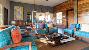 A spacious living area with a sofa, armchairs, and a coffee table. There is a kitchen in the background with a dining area.