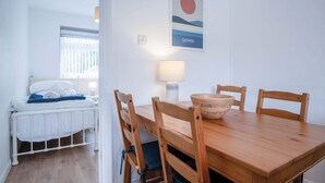 Una habitación con una mesa de comedor de madera, cuatro sillas, un cuenco de madera, una lámpara y una cama con ropa de cama blanca.