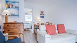 Un salón acogedor con un sofá blanco, un aparador de madera y un sillón azul. Hay una estantería con libros y una lámpara fijada a la pared.