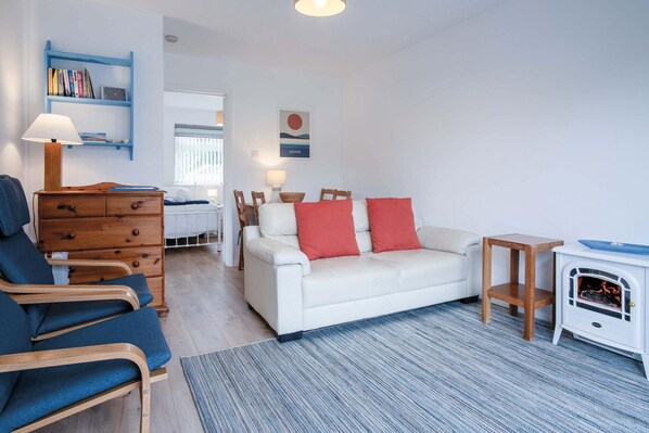 A modern living room with a white sofa, blue chairs, a wooden dresser, and a bookshelf. - Bayview 61 - 2 Bedroom Chalet - Bayview Oxwich (Swansea)