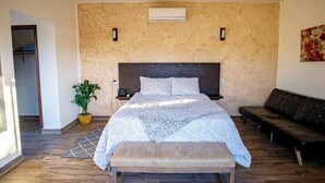 A bedroom with a large bed, a bench, a potted plant, a sofa, and an air conditioning unit on the wall.