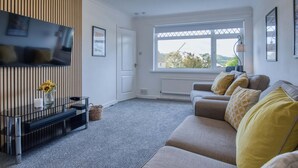 A modern living room with a grey sofa, a flat-screen TV mounted on the wall, and a large window providing a view of the outdoors.