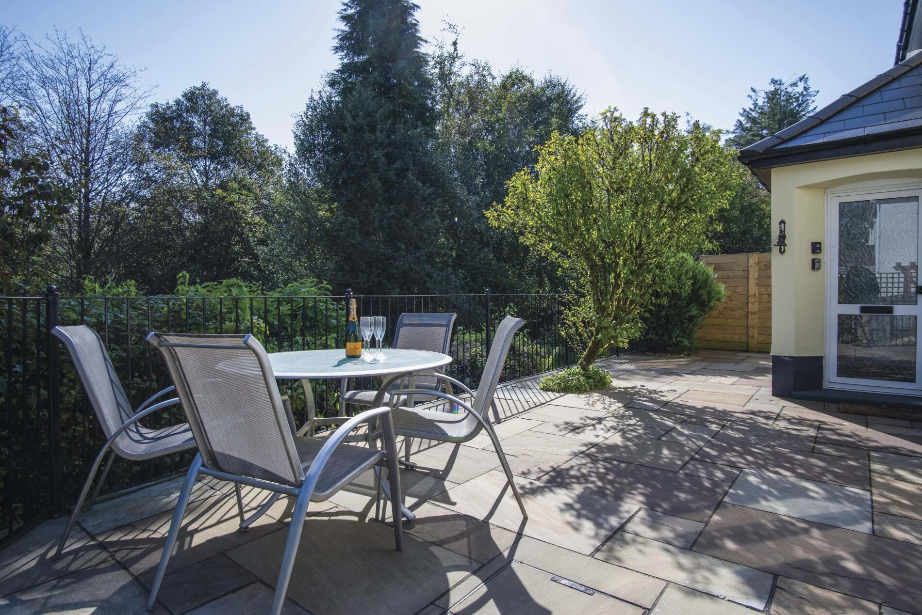 An outdoor patio with a round table and four chairs, surrounded by a metal fence and trees.