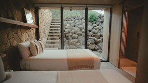 Een slaapkamer met een stenen muur, een bed met kussens, een houten plank en uitzicht op een rotsachtig landschap door een glazen deur.