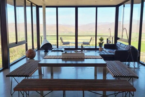A spacious room with a large table and benches, a sofa, and a view of the countryside through large windows.