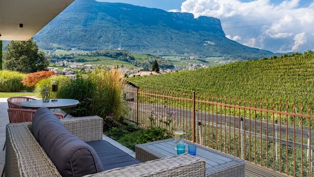 A terrace with wicker furniture, a table with a pitcher, and a view of a vineyard and mountains. - Apartment 'Landhaus Antonius Loft Suite Vitus' with Mountain View, Wi-Fi and Air Conditioning (St. Michael Eppan)