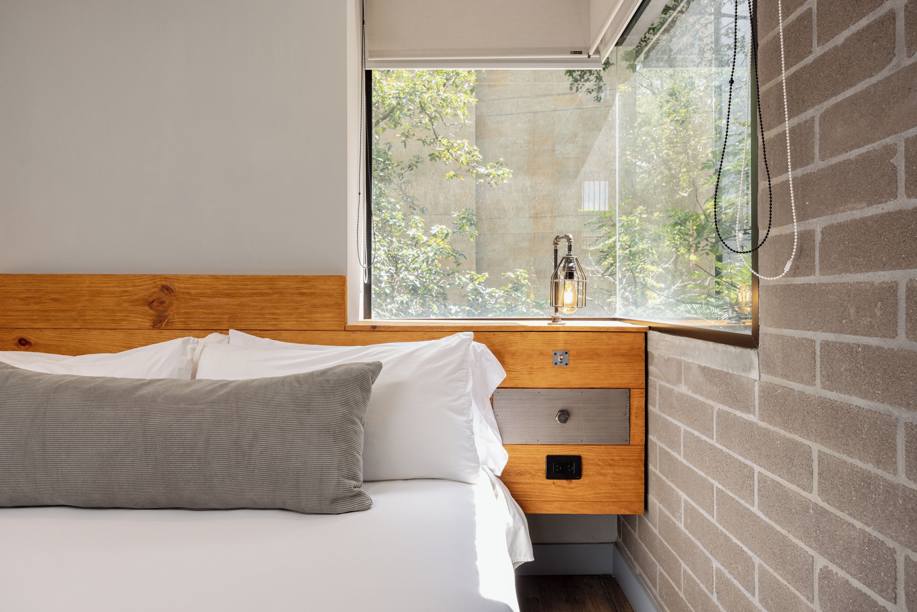 Une chambre avec une tête de lit en bois, un lit avec une literie blanche et grise, une table de chevet avec une lampe et une fenêtre donnant sur un paysage verdoyant.