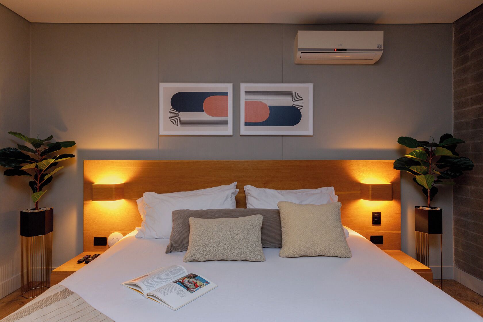 A modern hotel room with a wooden headboard, two bedside tables with lamps, a large bed with multiple pillows, and two wall-mounted air conditioning units.