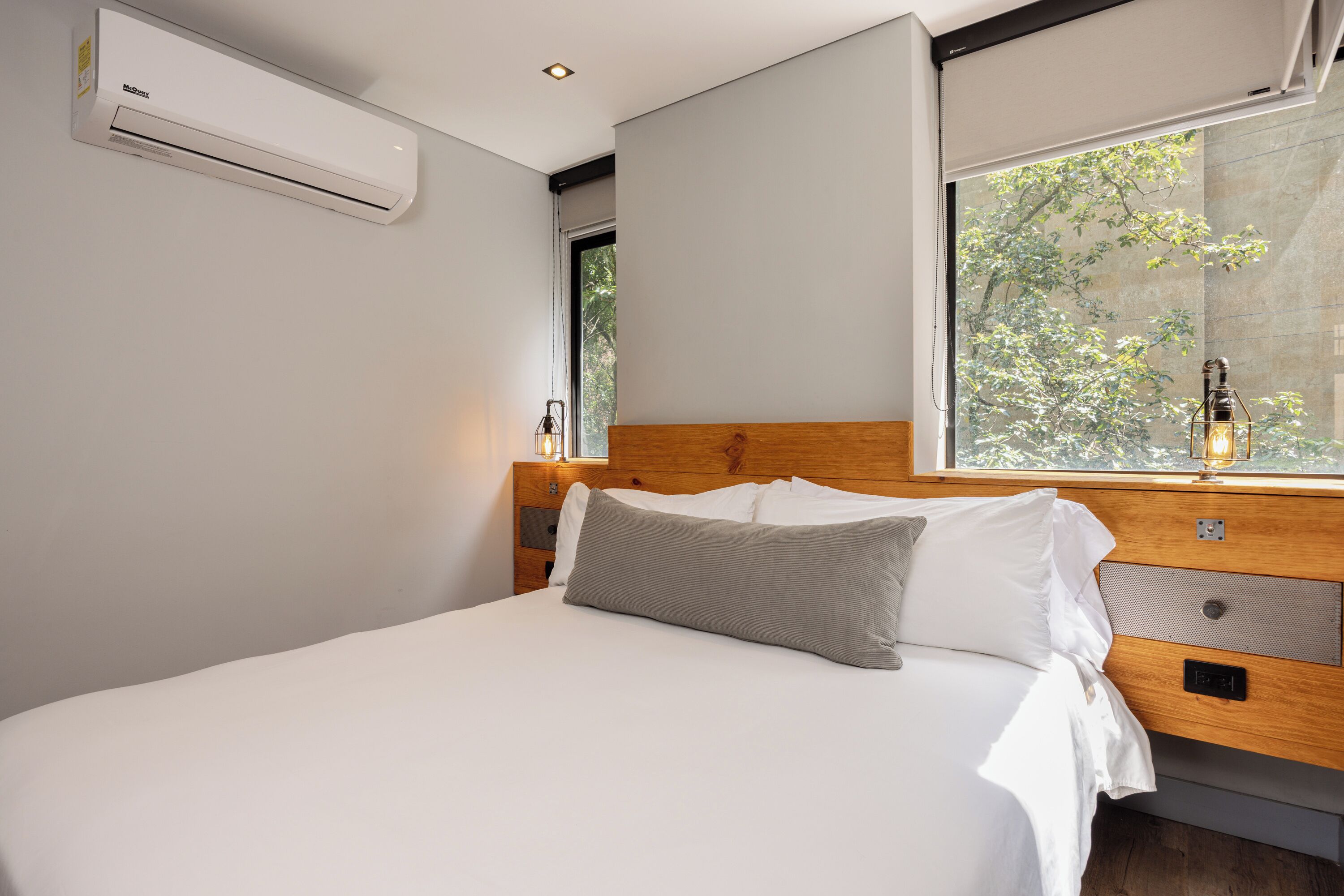 A modern bedroom with a large bed, a wooden headboard, a nightstand, and a window with a view of trees.