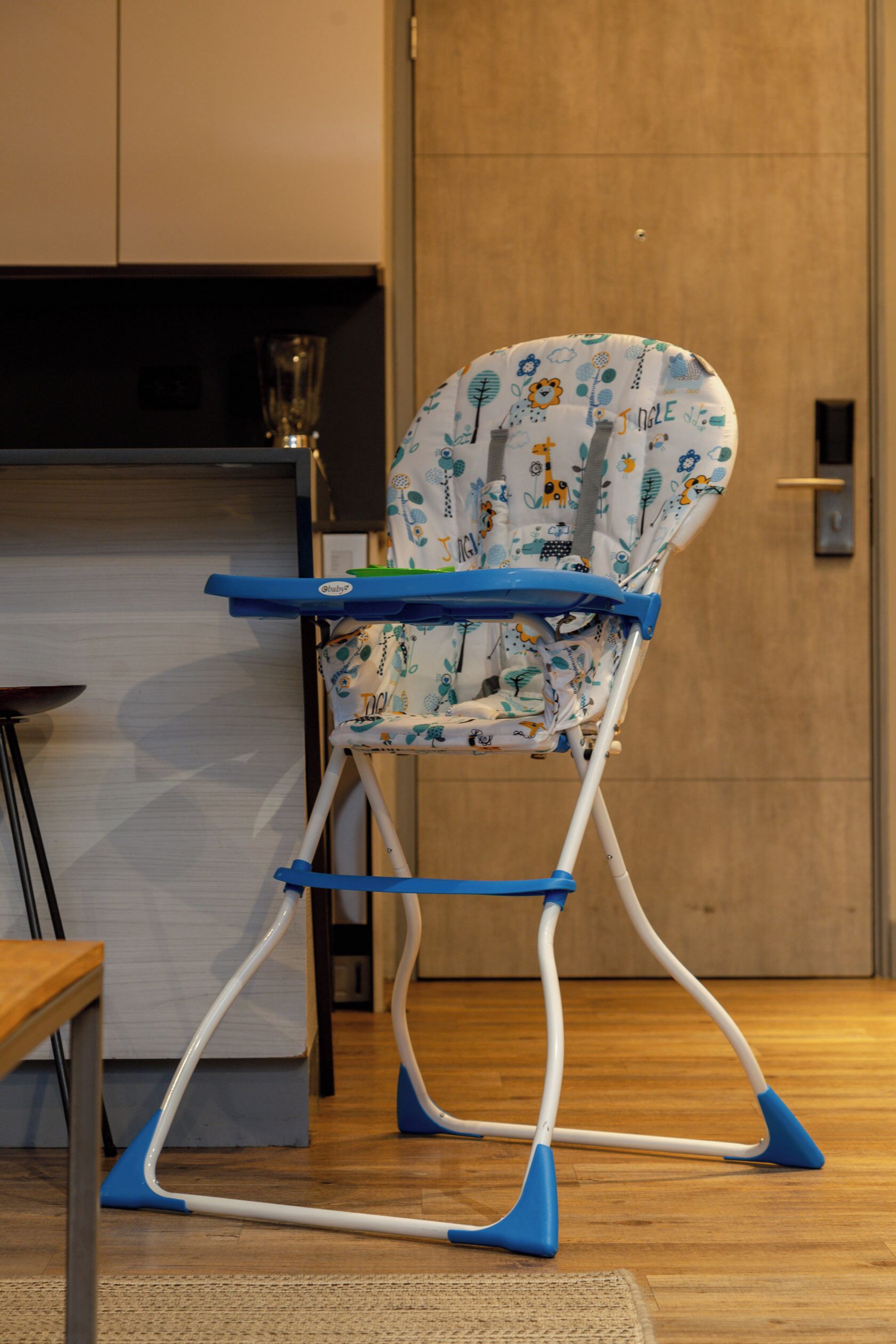 Une chaise haute avec un plateau bleu et un siège à motifs, dans une pièce dont le sol est en bois et où l’on aperçoit une porte au fond.