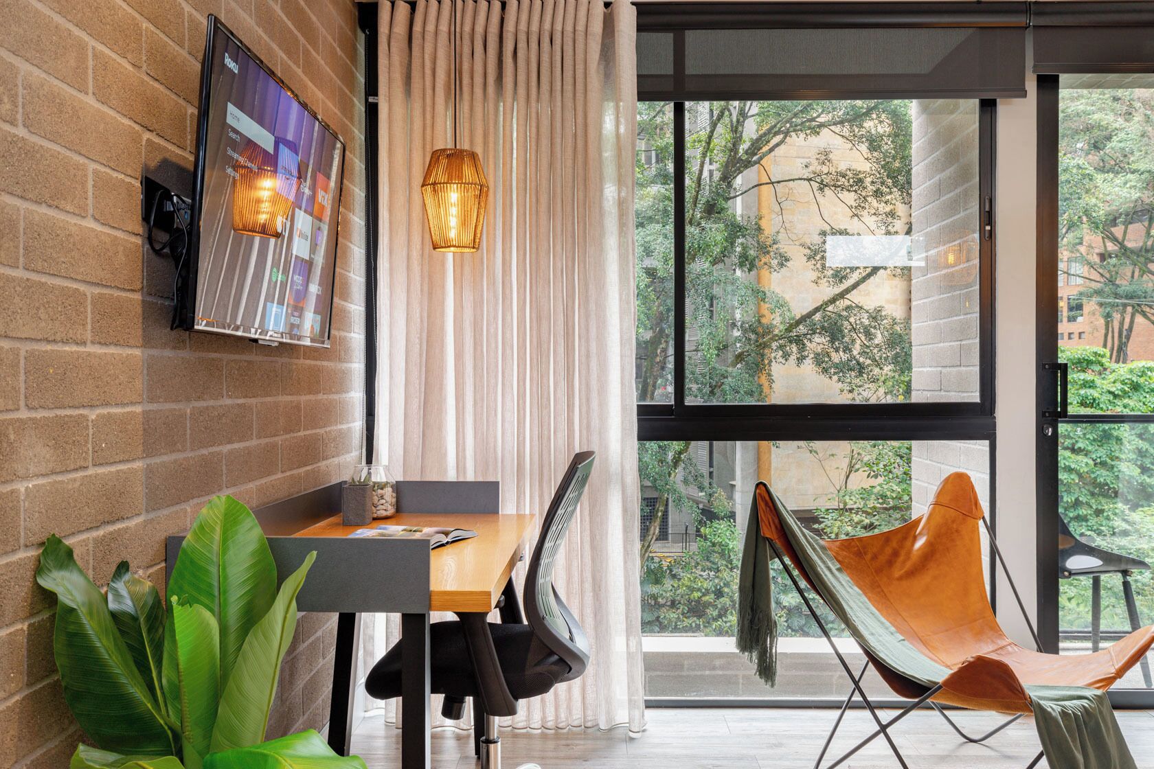 A modern dining area with a brick wall, a TV mounted on the wall, a table with chairs, and a view of greenery outside.