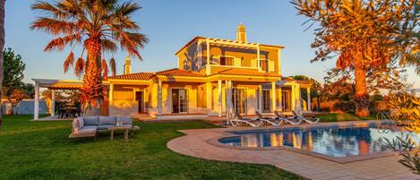 Una casa de dos pisos con piscina, un patio con sillones de descanso y un césped bien cuidado.