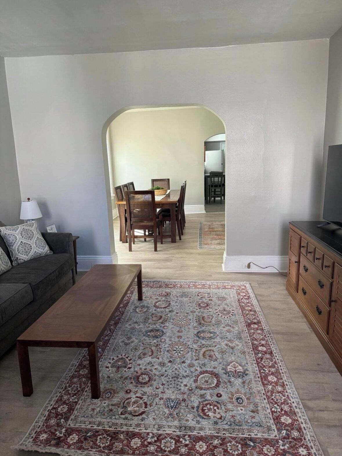 Una sala de estar con un sofá oscuro, una mesa de centro de madera y un tapete estampado. Se observa una zona de comedor con sillas de madera y una mesa al fondo.