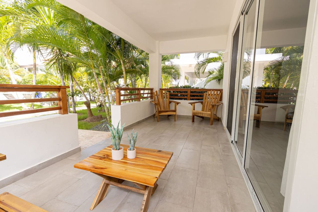 Ein überdachter Patio-Bereich mit Holzmöbeln, Blumentöpfen und Blick auf Palmen.