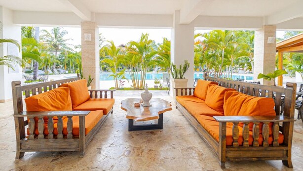 Un espace détente avec deux canapés en bois dotés de coussins orange, une table basse centrale et une vue sur une piscine et des palmiers.