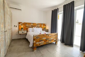 A bedroom with a wooden headboard, a bed with white bedding, a bedside table with a lamp, and a window with curtains. - Coastal Luxury 2-Bed w Pool Access in Bayahibe (Bayahíbe)