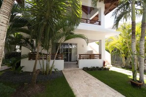 A modern house with a covered patio, palm trees, and a stone pathway leading to the entrance. - Coastal Luxury 2-Bed w Pool Access in Bayahibe (Bayahíbe)