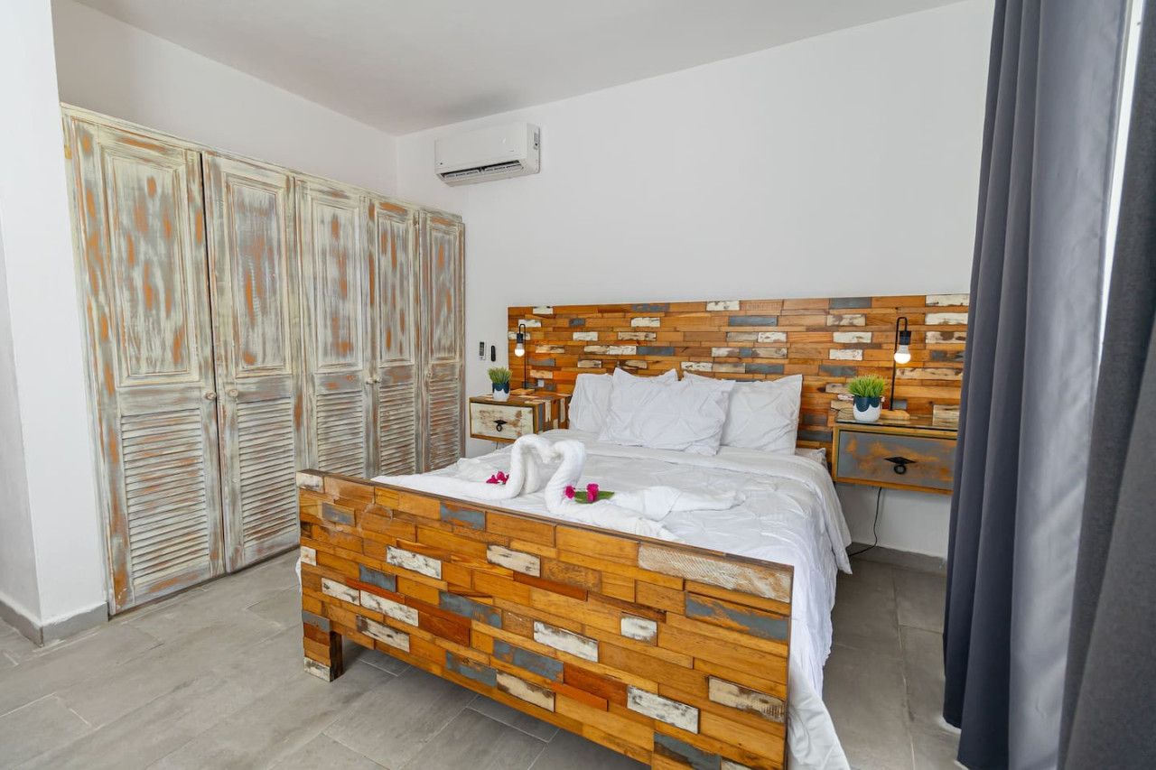 Een slaapkamer met een houten hoofdbord, een bed met witte lakens, twee nachtkastjes, een kleine plant in een pot en een kledingkast met een rustieke afwerking.