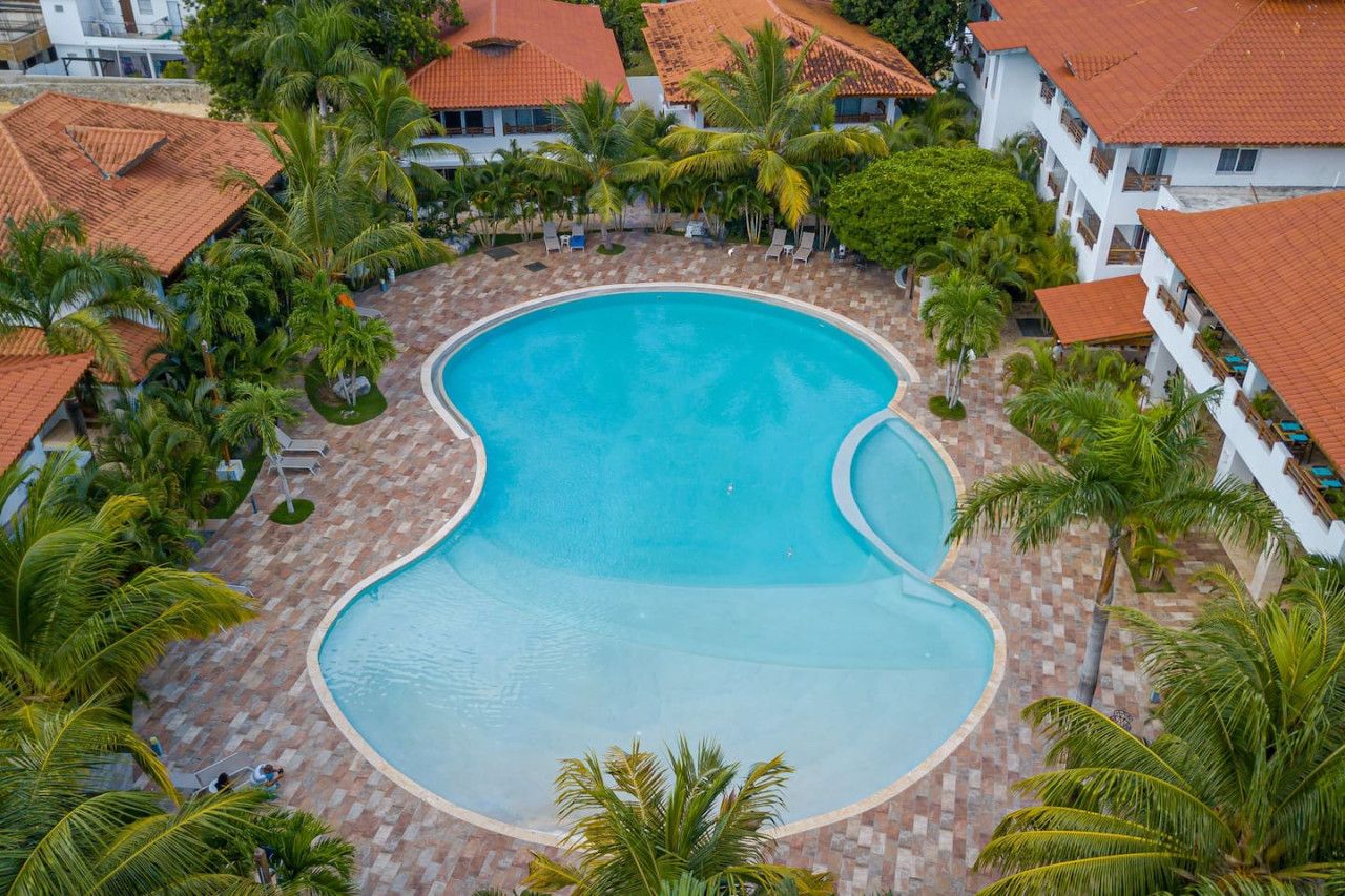 An aerial view of a resort with a large swimming pool surrounded by palm trees and buildings with red-tiled roofs.