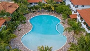 An aerial view of a resort with a large swimming pool surrounded by palm trees and buildings with red-tiled roofs.
