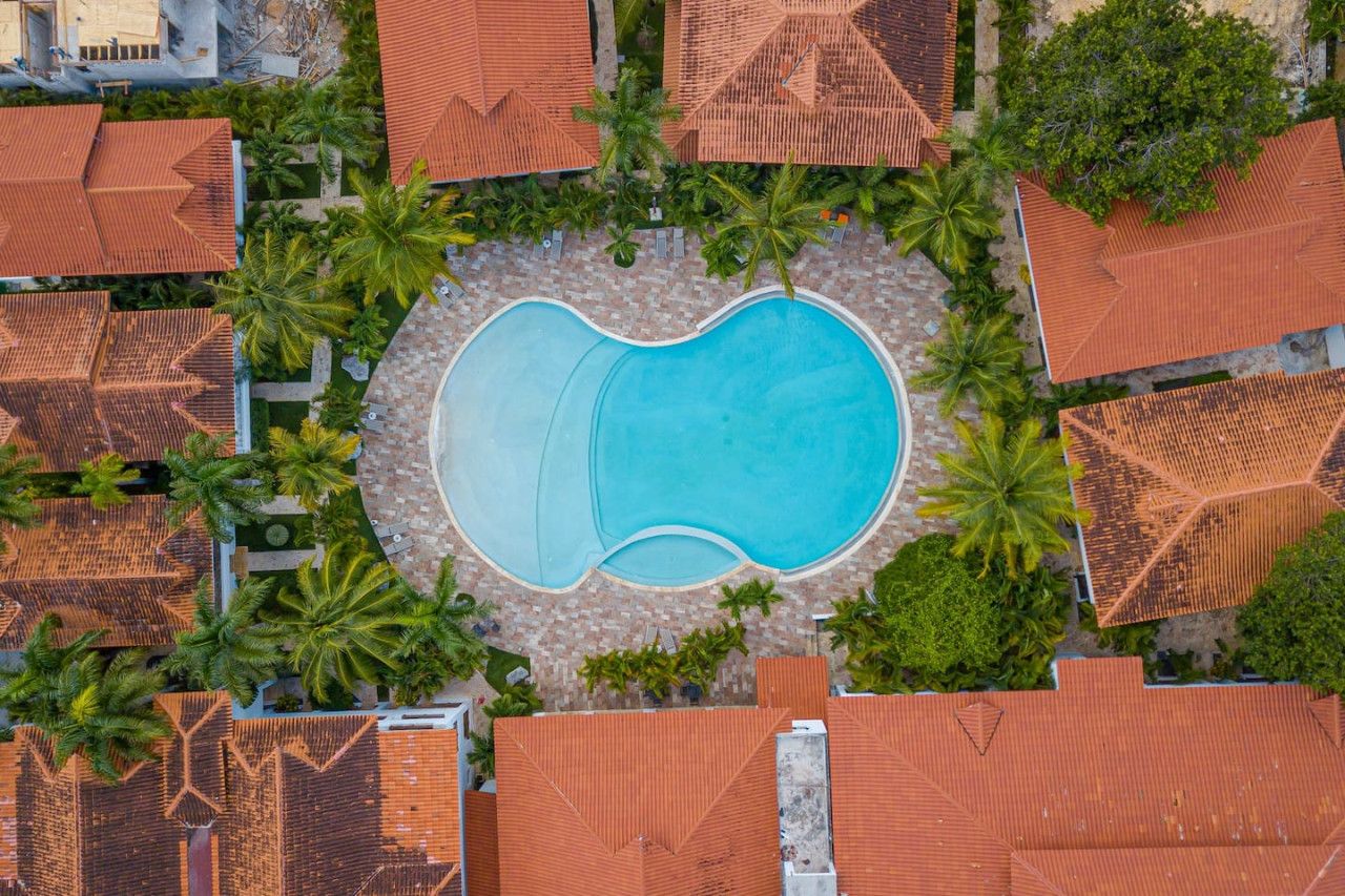 Eine Vogelperspektive zeigt ein Resort mit einem zentralen Schwimmbecken, umgeben von Palmen und roten Ziegeldächern.