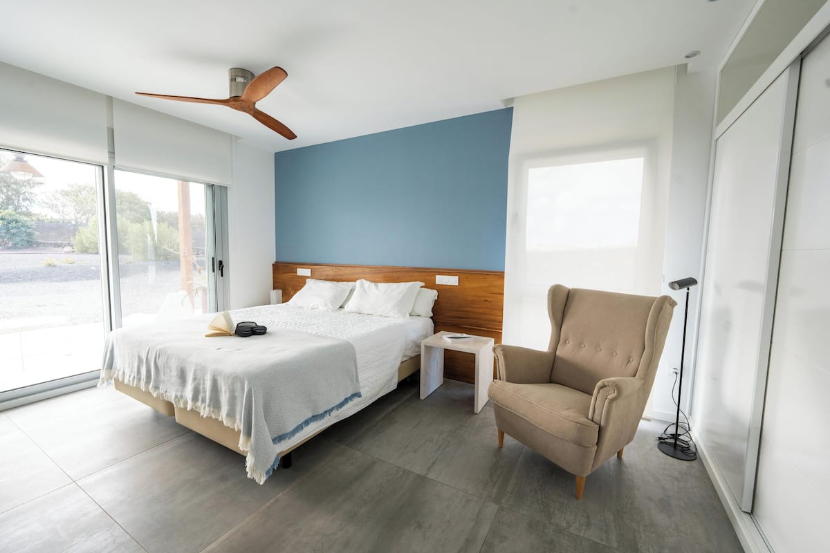 Un dormitorio moderno con una cama grande, un cabecero de madera, un sillón color beige, una mesita de noche y una lámpara de pie.