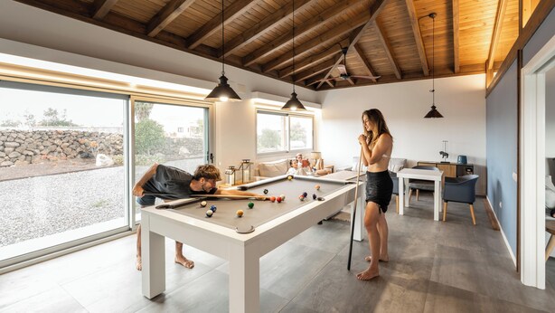 Una mesa de billar moderna en un interior, con una pared de piedra y un amplio ventanal al fondo.