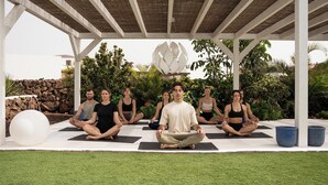 Un grupo de personas meditando en un espacio al aire libre techado, con una pared de piedra y follaje en el fondo.