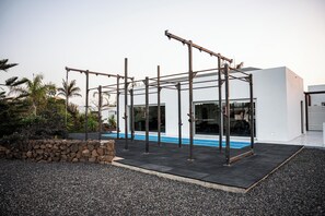 A modern outdoor gym with metal frames and rubber flooring, set against a backdrop of a white building and a stone wall. - Kyuka Surfclub (La Oliva)