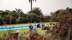 Un grupo de personas practicando yoga al aire libre cerca de una piscina, rodeado de exuberante follaje y un muro de piedra.